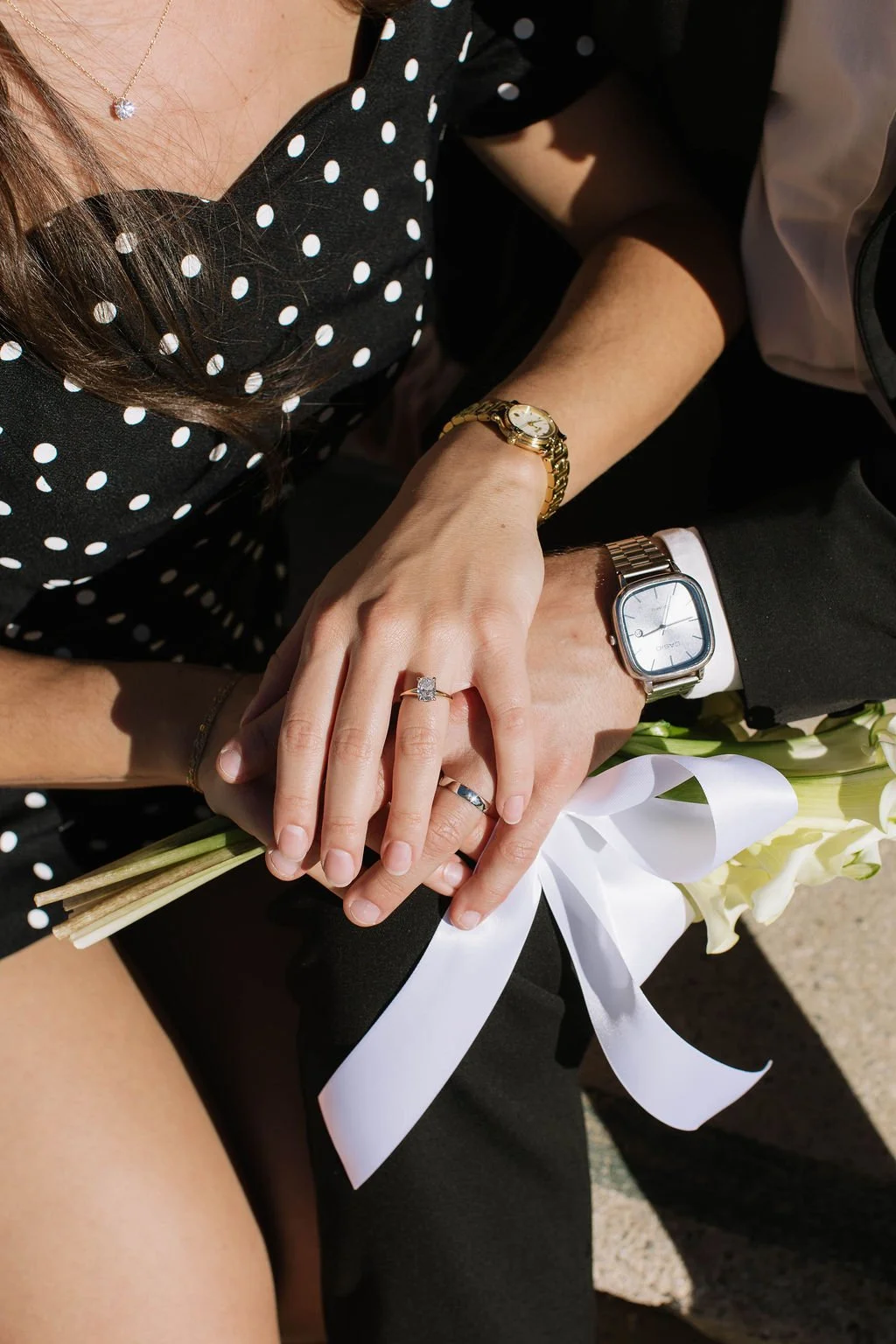 Close-up of a couple holding hands, showing wedding rings, watches, and a bouquet of white flowers with a white ribbon.