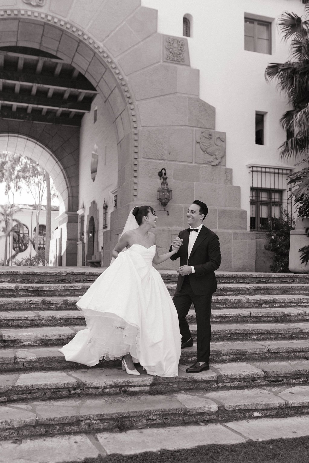 A black and white photo of a bride and groom dancing on stone steps outside a historic building with ornate architecture, arched doorway, and palm trees.