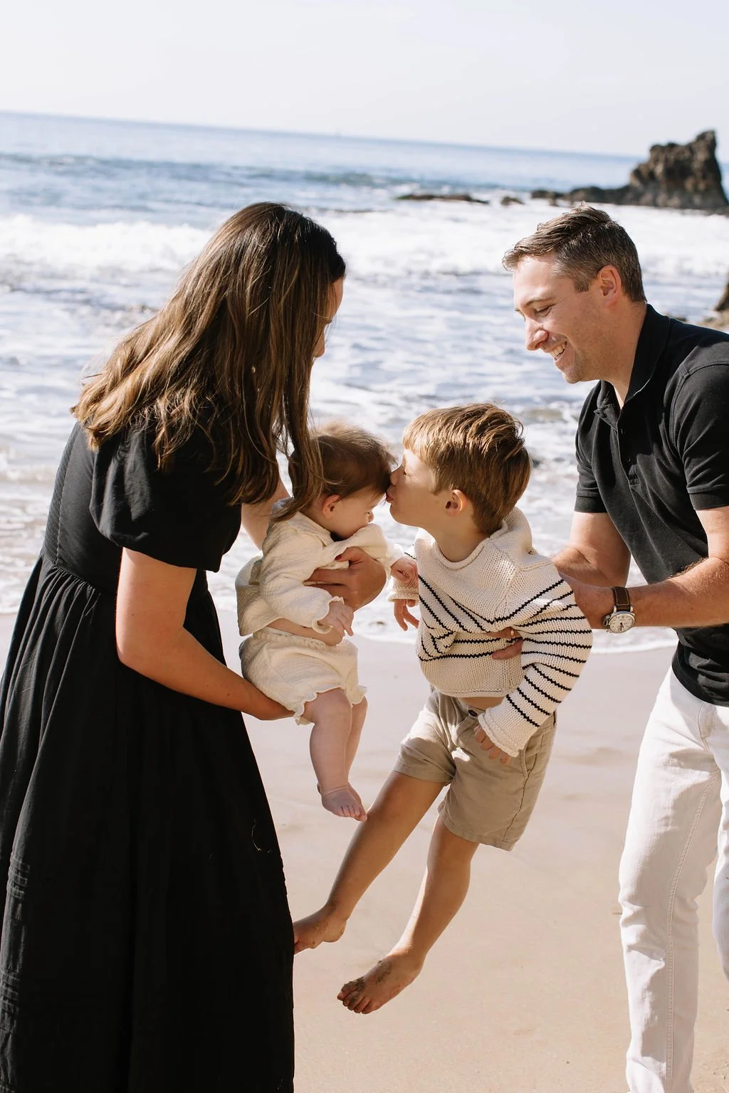 Family on the beach, with parents holding and kissing their children, one of the children is a baby, the other is a young boy.