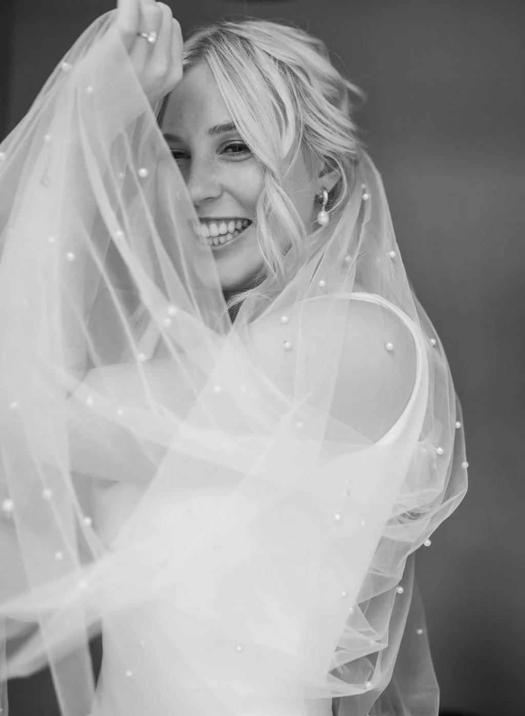 Black and white photo of a smiling woman with wavy blonde hair, partially covered by a sheer veil with small pearl embellishments.
