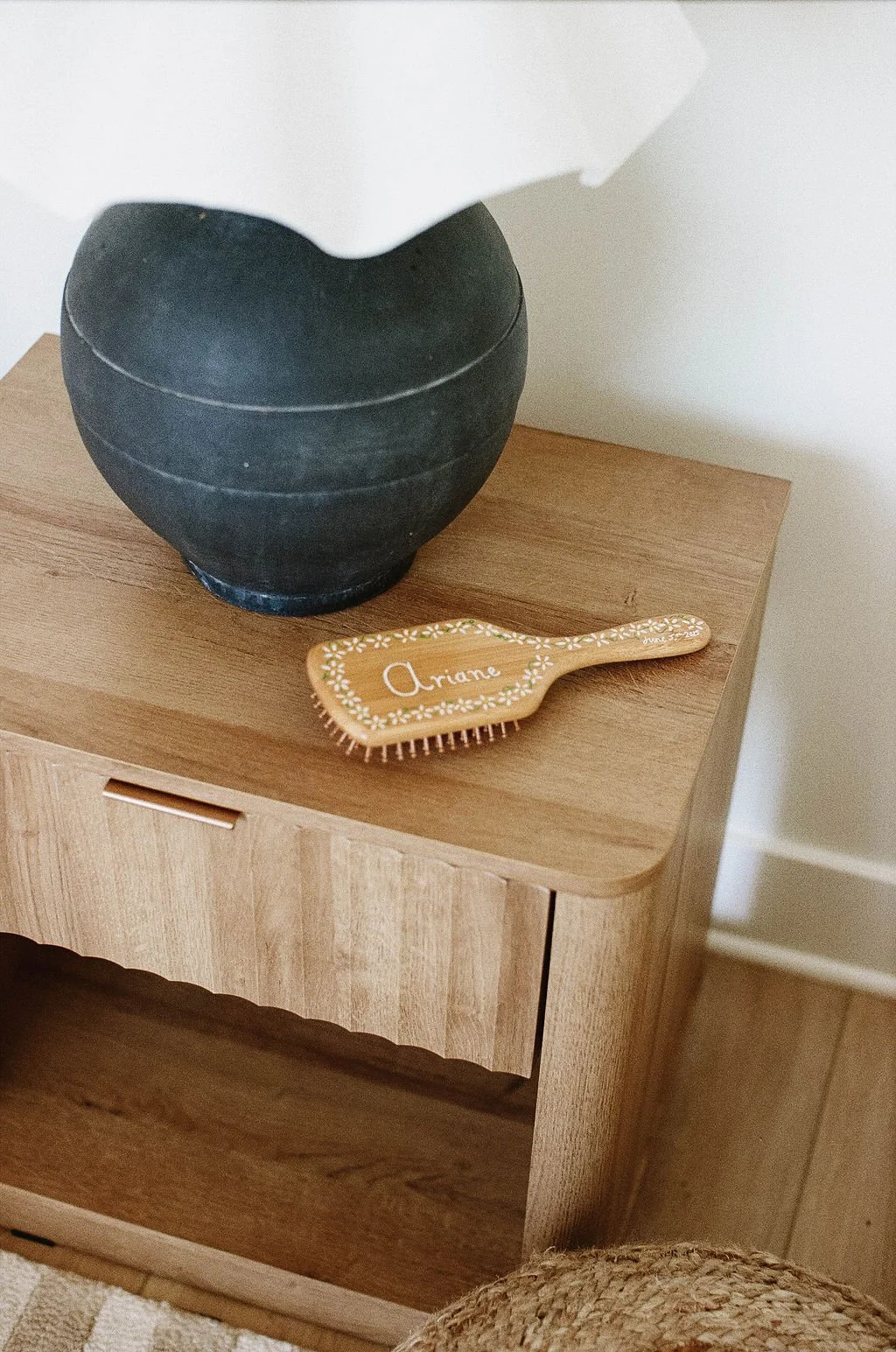 A black round vase and a wooden hairbrush with the name 'Arianne' on it are placed on a wooden side table.