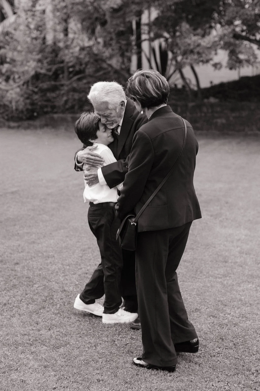A black and white photo of three people, two women and a young boy, hugging outdoors on a grassy area with trees in the background. The older woman has gray hair and wears glasses, the younger woman has dark hair, and the boy has dark hair and is wearing a light-colored shirt and dark pants.