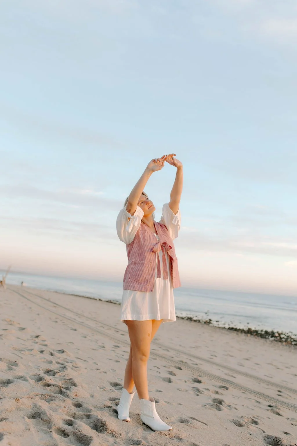 A woman standing on a sandy beach during sunset, wearing a pink vest, white dress, and white boots, holding a seashell above her head, with the ocean and cloudy sky in the background.
