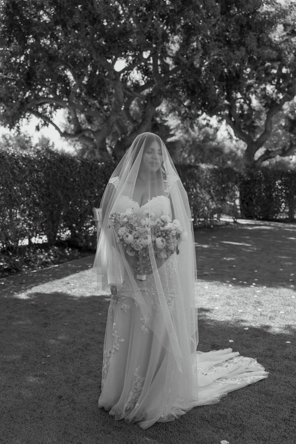 A bride in a wedding dress holding a bouquet of flowers, wearing a veil, standing outdoors with trees and bushes in the background.
