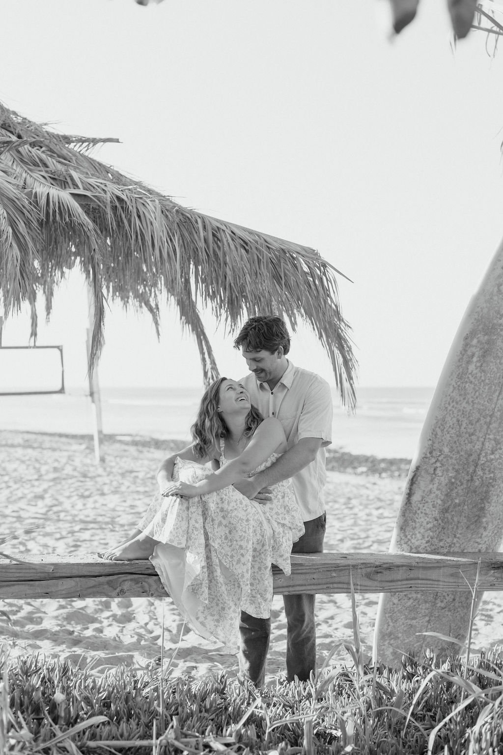A black and white photo of a young couple smiling and holding each other on a beach under a thatched-roof hut, with a surfboard leaning against a post nearby.