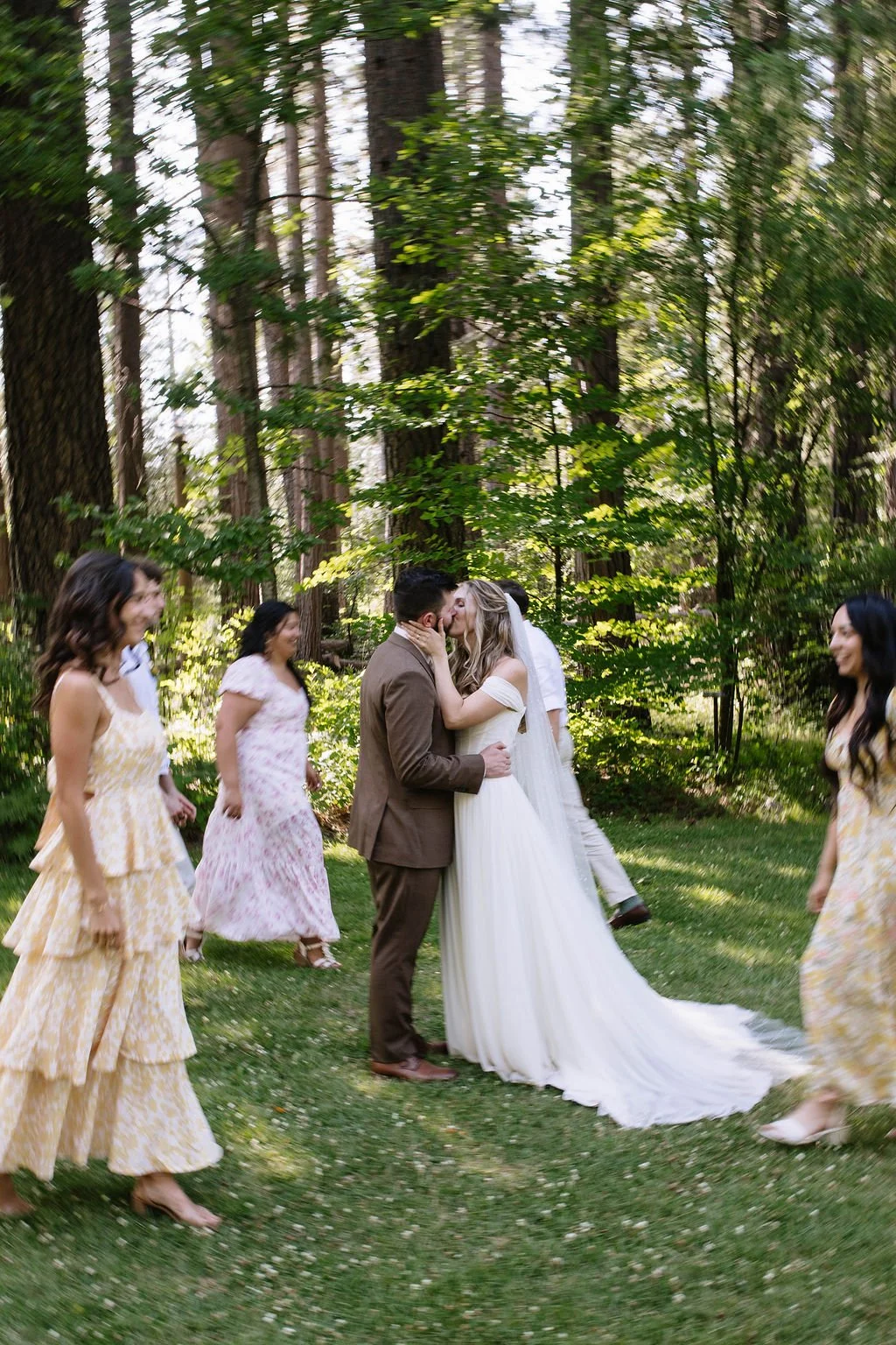 A wedding ceremony in a wooded outdoor setting, with a bride and groom kissing, surrounded by bridesmaids and possibly groomsmen, all dressed in formal attire.