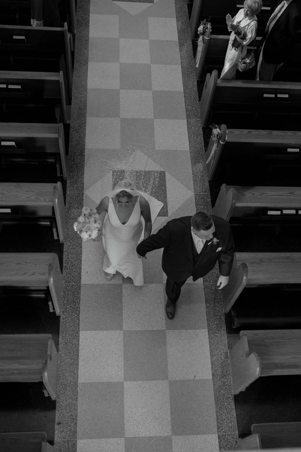 Black and white photo of a wedding ceremony inside a church. A bride in a white gown with a bouquet is walking down the aisle, with a groom in a dark suit walking beside her. An older woman in a light-colored dress and glasses is standing in the pews, clapping.
