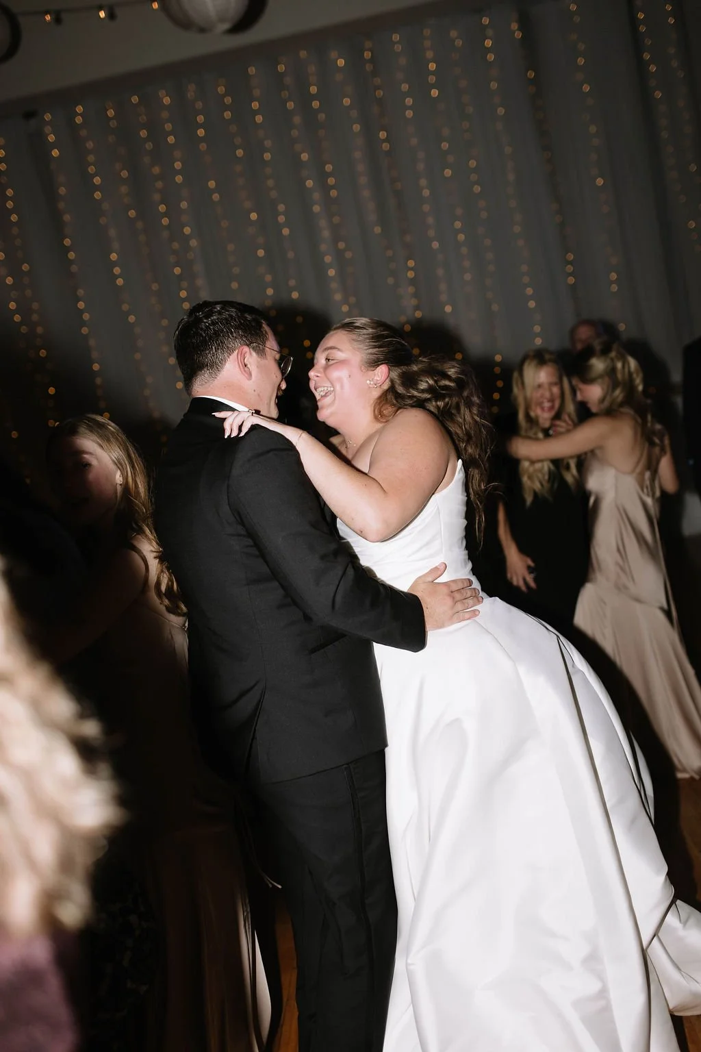 A bride and groom are dancing happily at their wedding, surrounded by guests, with warm lighting and string lights in the background.