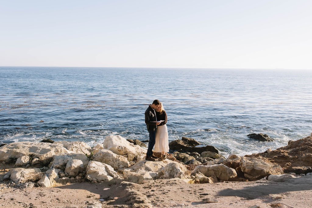A couple stands on rocky beach near the ocean, embracing and reading a book.