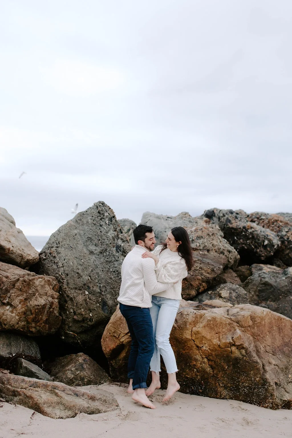A couple laughing and embracing on a sandy beach surrounded by large rocks, with seagulls flying in the cloudy sky.