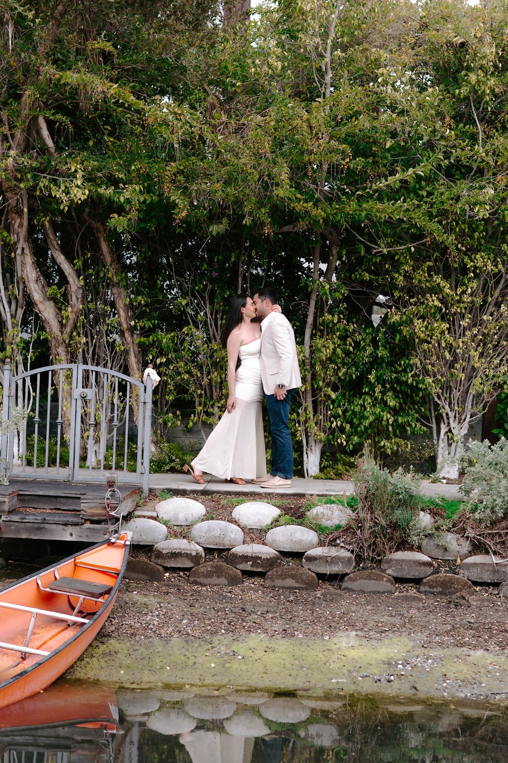 A couple sharing a kiss on a small wooden dock by a pond, surrounded by lush trees, with an orange canoe in the water.