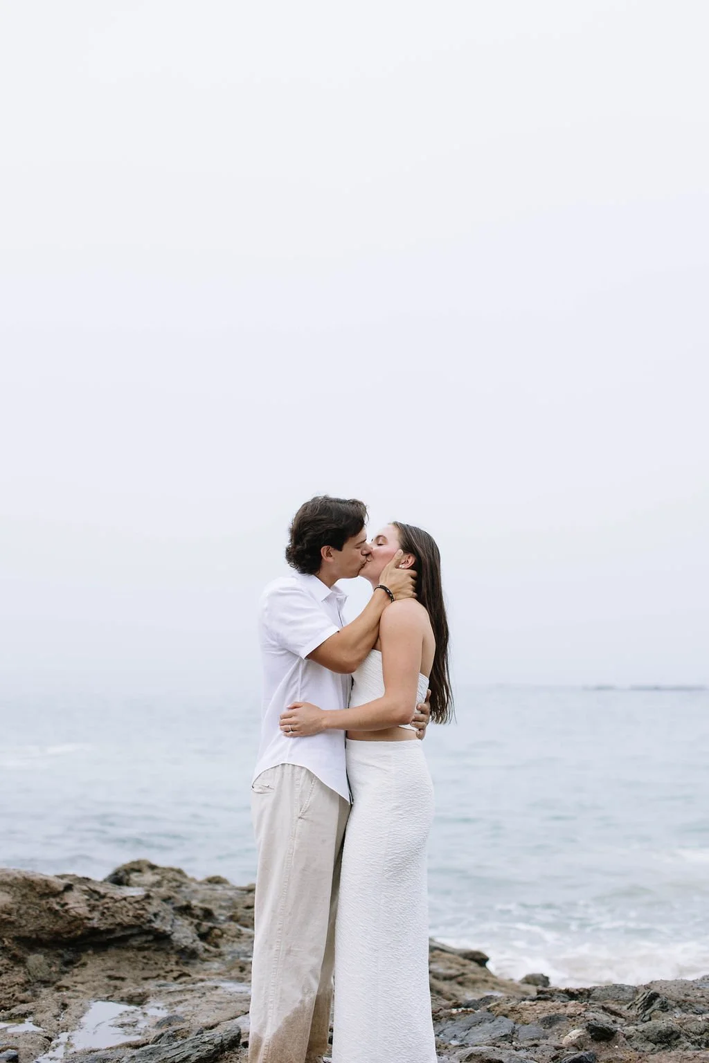 A young couple sharing a kiss on a rocky beach with the ocean in the background on a cloudy day.