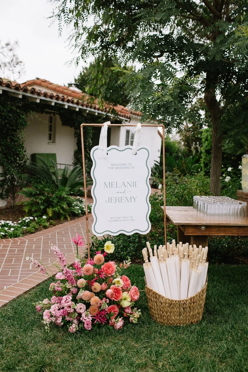 Outdoor wedding sign with floral decoration, welcoming guests to Melanie and Jeremy's wedding, placed next to a basket of paper straws and glass cups, on a grassy area with a brick pathway, greenery, and trees in the background.