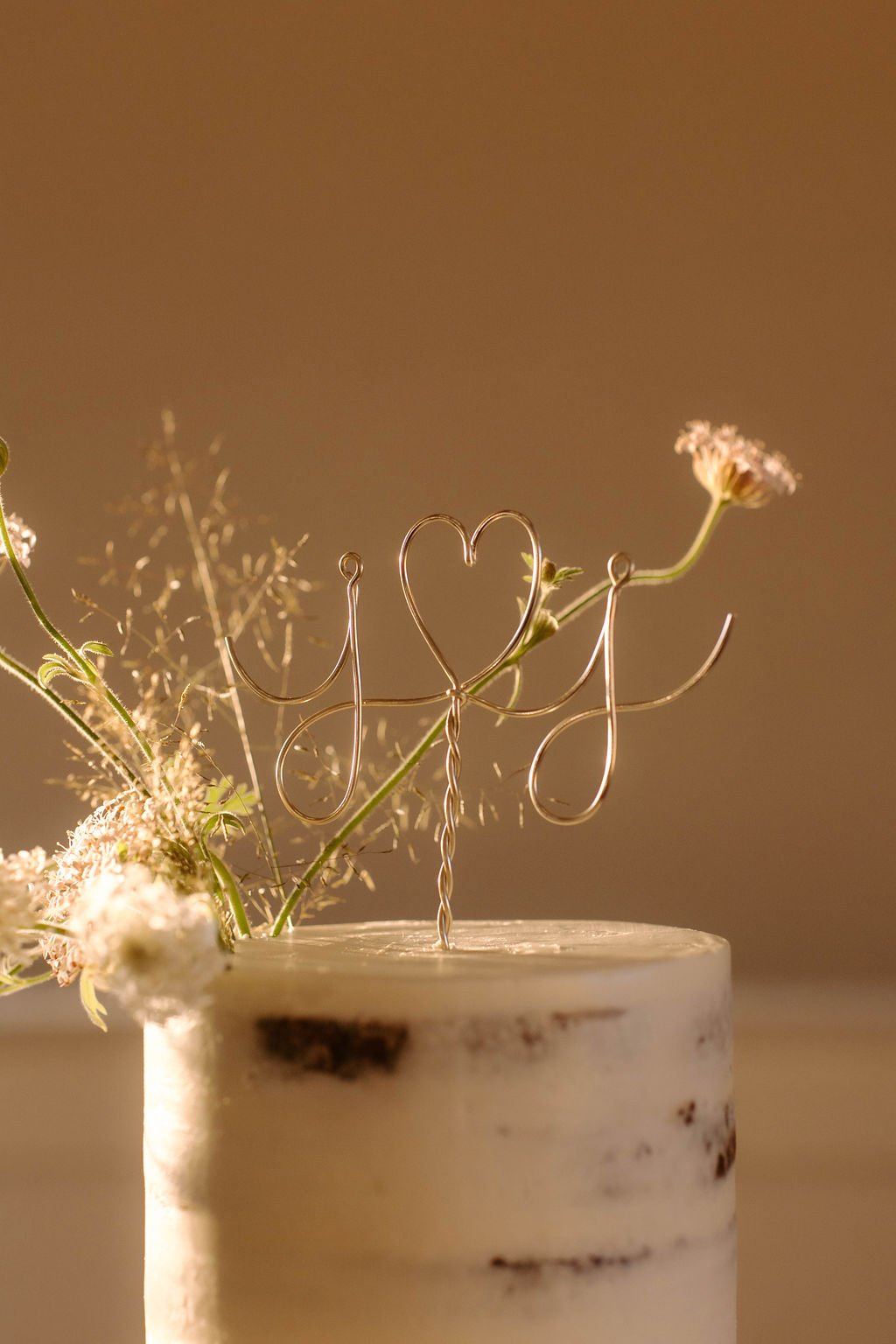 A white cake with a wire topper spelling 'joy' with a heart in place of the letter 'o', decorated with small flowers and greenery, against a warm background.