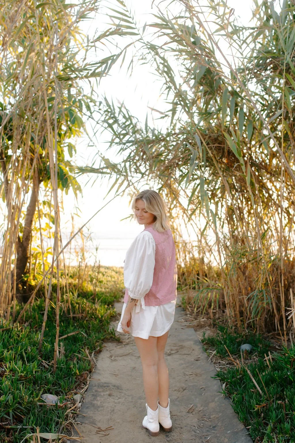 A woman walking on a sandy path surrounded by tall green and brown plants, with the sunset in the background.