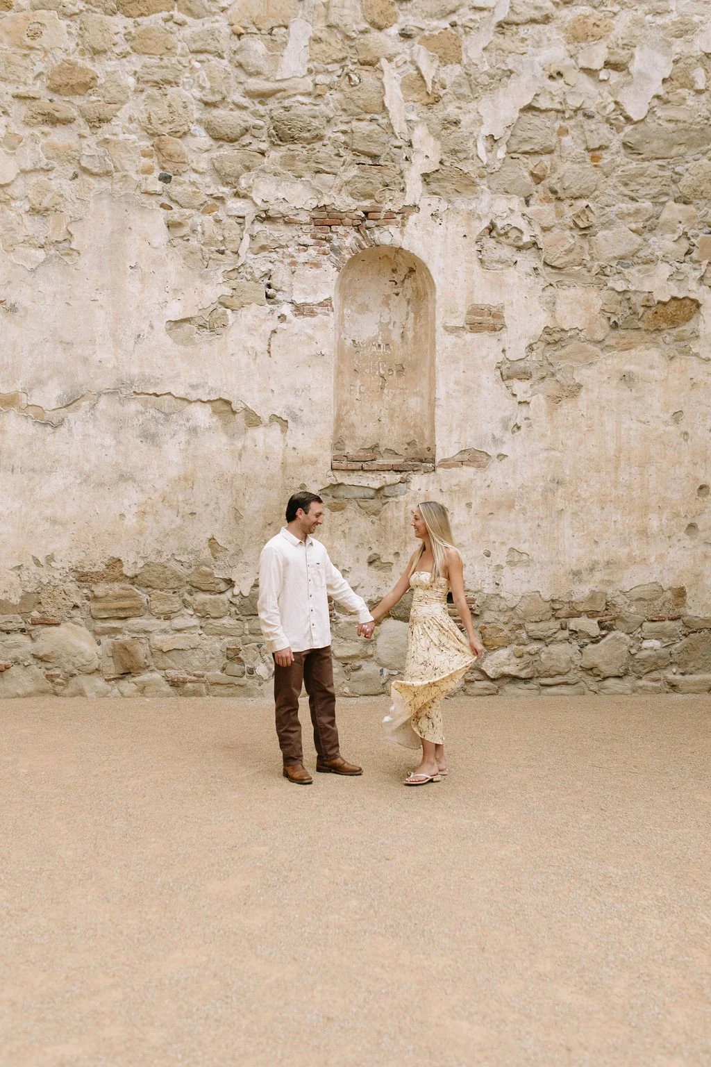 A man and woman standing and holding hands in front of a weathered stone wall, with the woman lifting her dress slightly as they smile at each other.