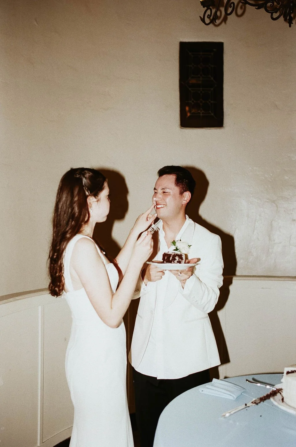A man and woman, dressed in wedding attire, sharing a moment with the groom holding a plate with a piece of cake, while the bride playfully touches the groom's nose during a wedding celebration.