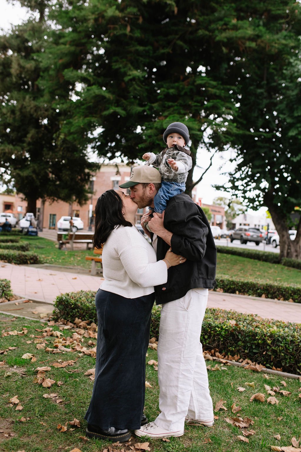 A couple sharing a kiss while a young child sits on the man's shoulders in a park with trees and buildings in the background