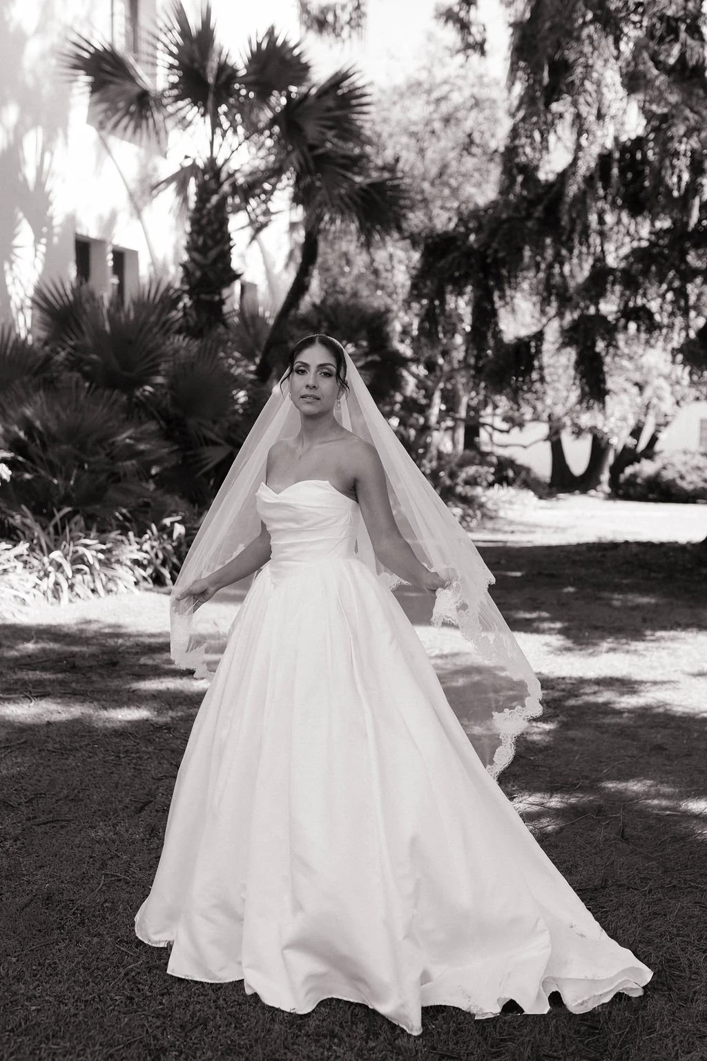 Black and white photo of a bride in a strapless wedding gown and veil outdoors in a garden with trees and plants.