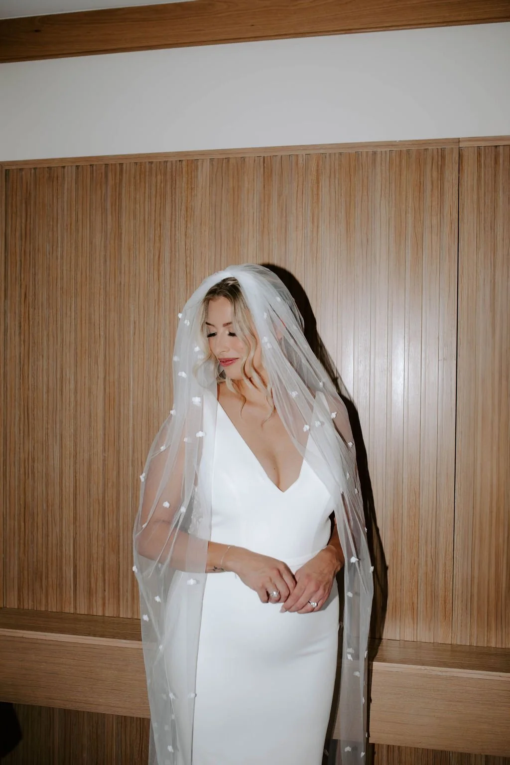 Bride in a white wedding dress with a veil, standing in front of a wooden wall, looking down with a gentle smile.