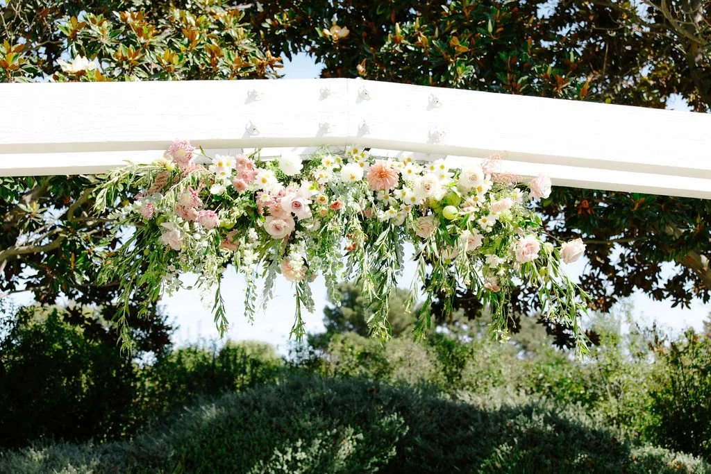 Flower arrangement with pink and white flowers hanging beneath a white structure outdoors, with trees in the background.