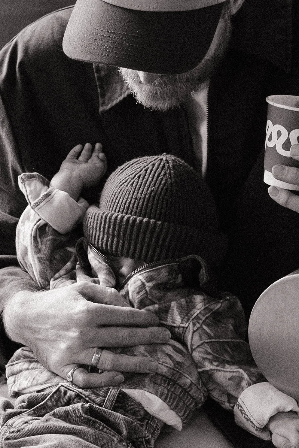 Black and white photo of a person holding a sleeping baby wrapped in a jacket and beanie, with one hand resting on the baby's back and another holding a soda cup.