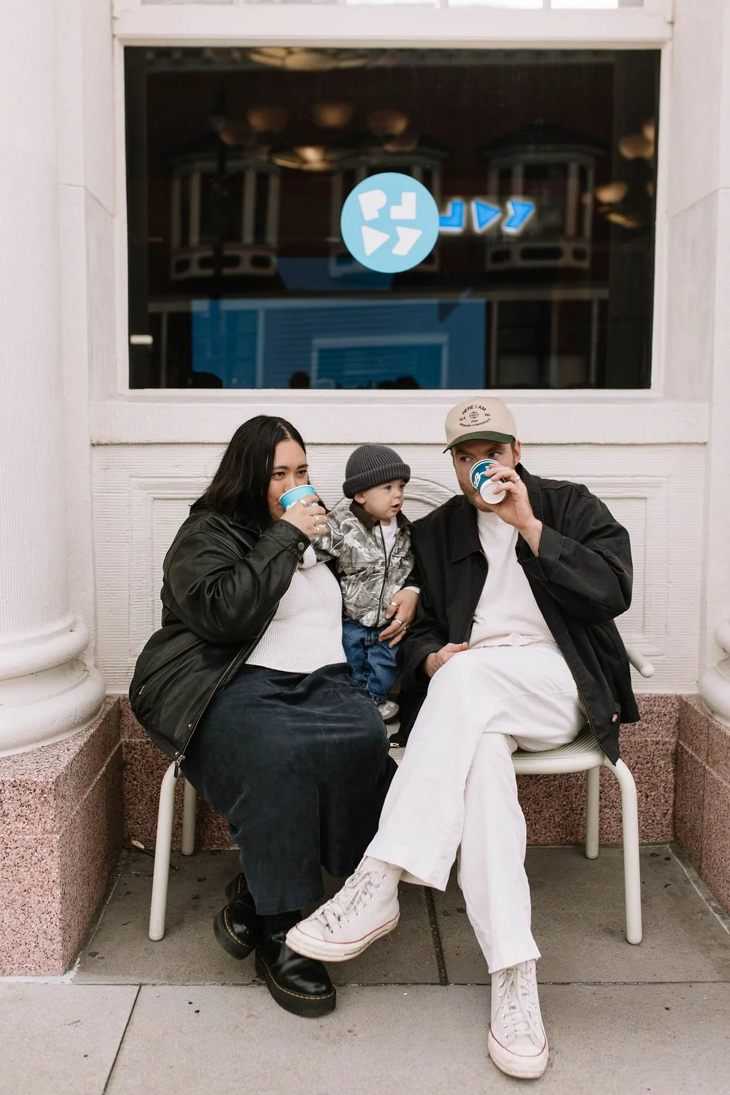 A family of three sitting on a bench outside, drinking coffee or hot chocolate. The woman is wearing a black jacket and a black skirt, the man is wearing a black jacket, white pants and a cap, and the child, standing between them, is wearing a gray beanie and camouflage jacket.
