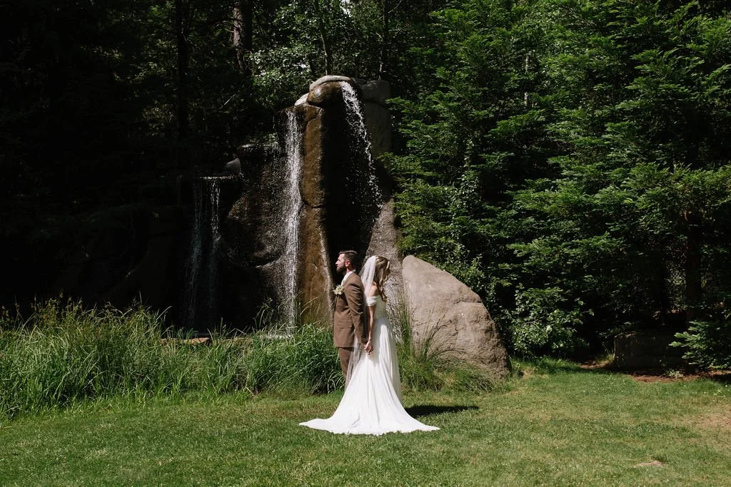 A bride and groom stand back-to-back outdoors near a waterfall and lush green trees.