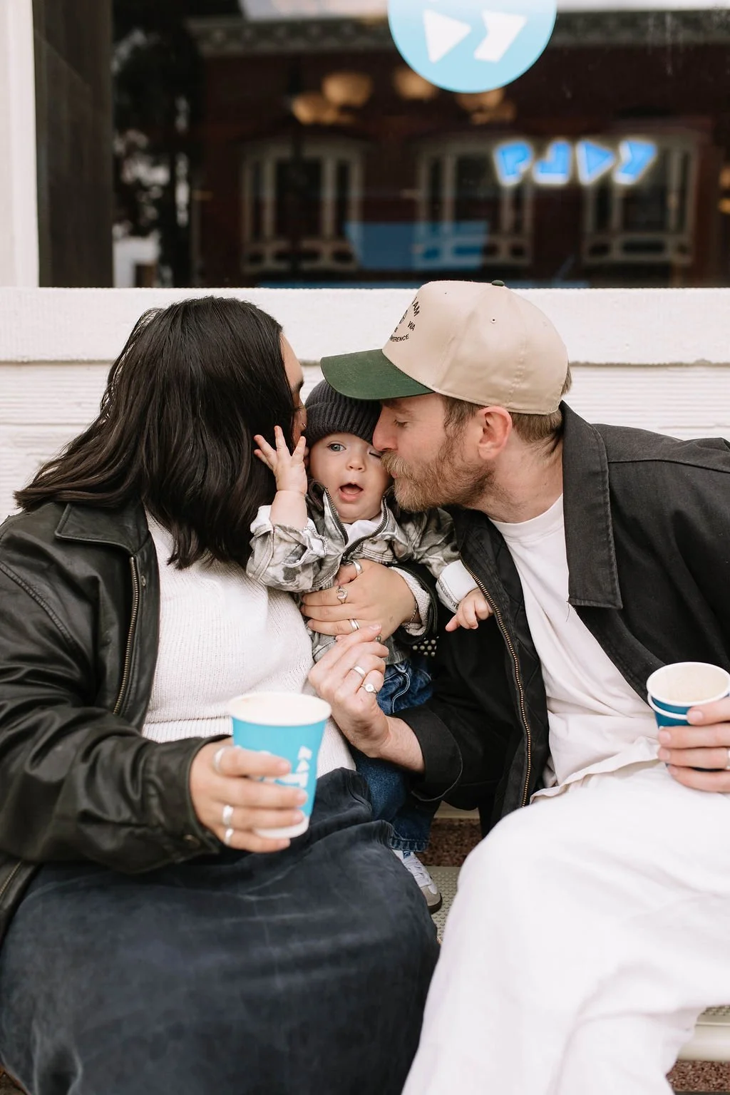 A family of three sitting on a bench outdoors, sharing a kiss with their young child between them, holding cups as they celebrate.