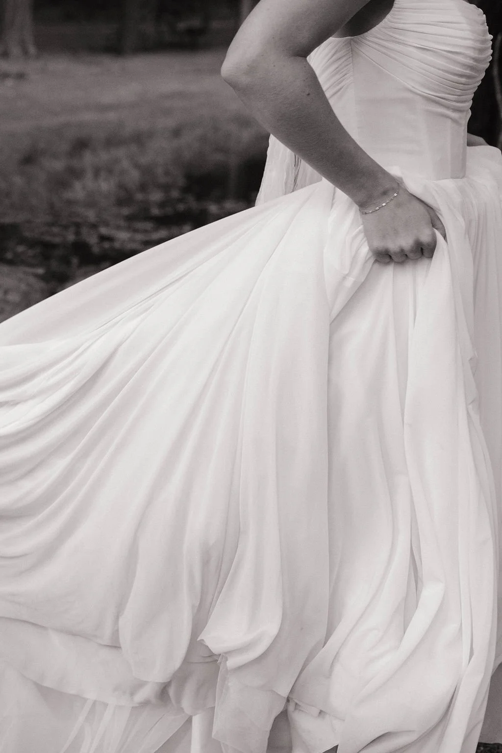 Close-up of a woman wearing a white wedding dress, holding the fabric of her gown with one hand, outdoors setting in black and white.