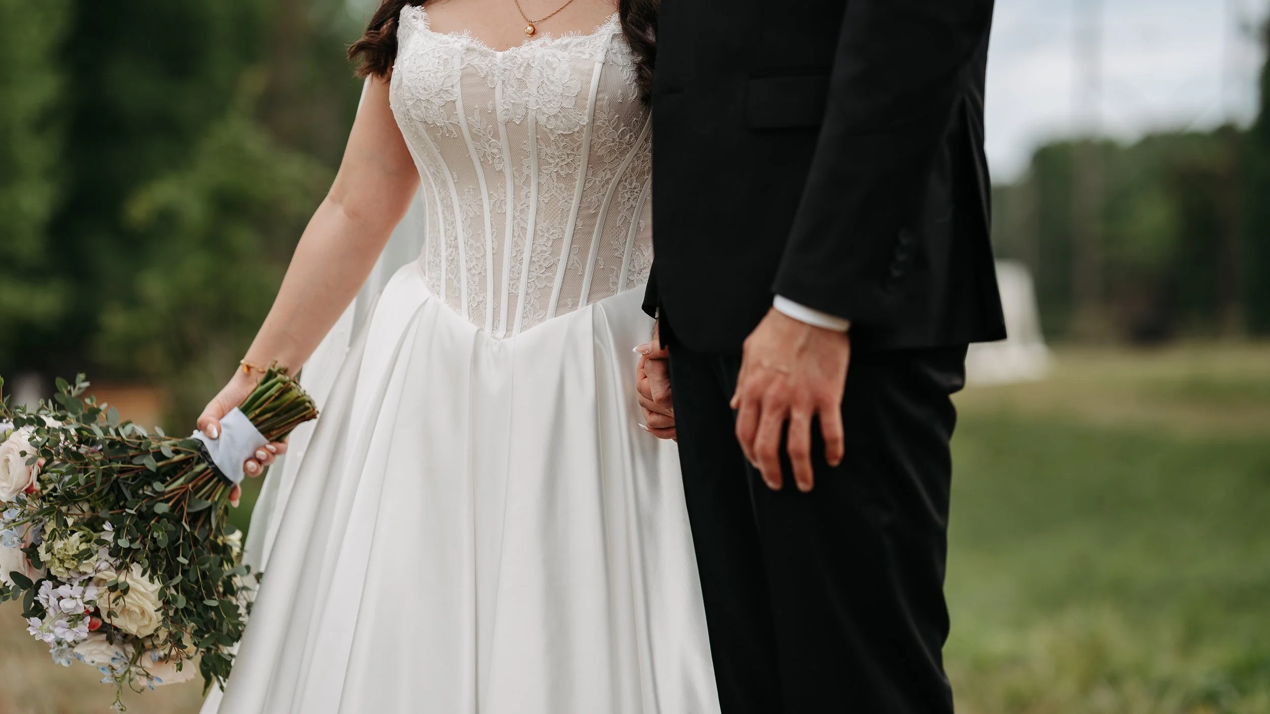 Close-up of a bride and groom holding hands at their wedding, with the bride in a lace dress holding a bouquet of flowers, and the groom in a black suit, outdoors during the daytime. Kristen Dianne Photo in Columbia, SC Photographer Weddings
