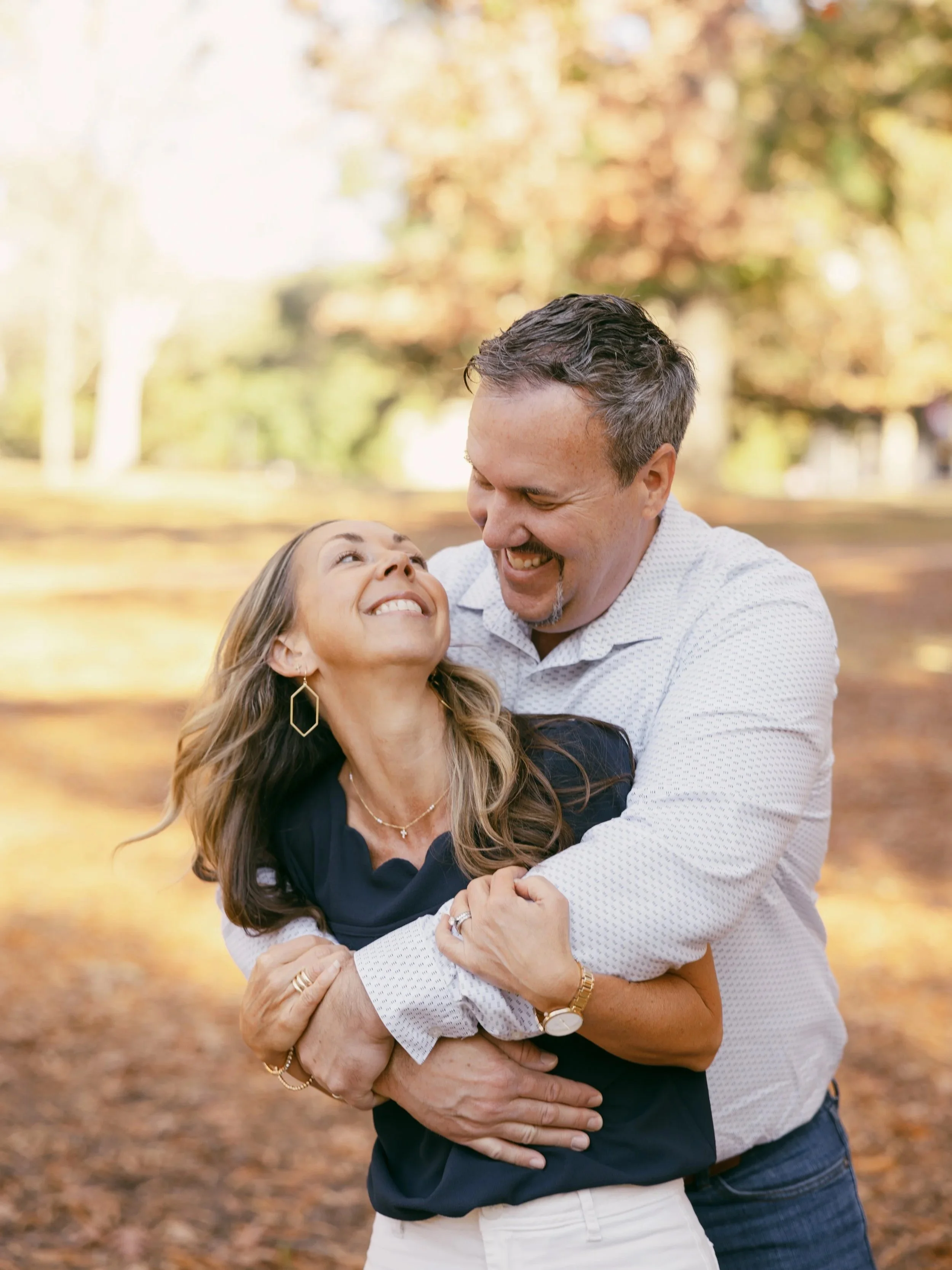 A happy couple at the Horseshoe during autumn, holding each other and smiling, with fallen leaves on the ground and trees with yellow and orange foliage in the background. Kristen Dianne Photo in Columbia, South Carolina. SC Photographer Family