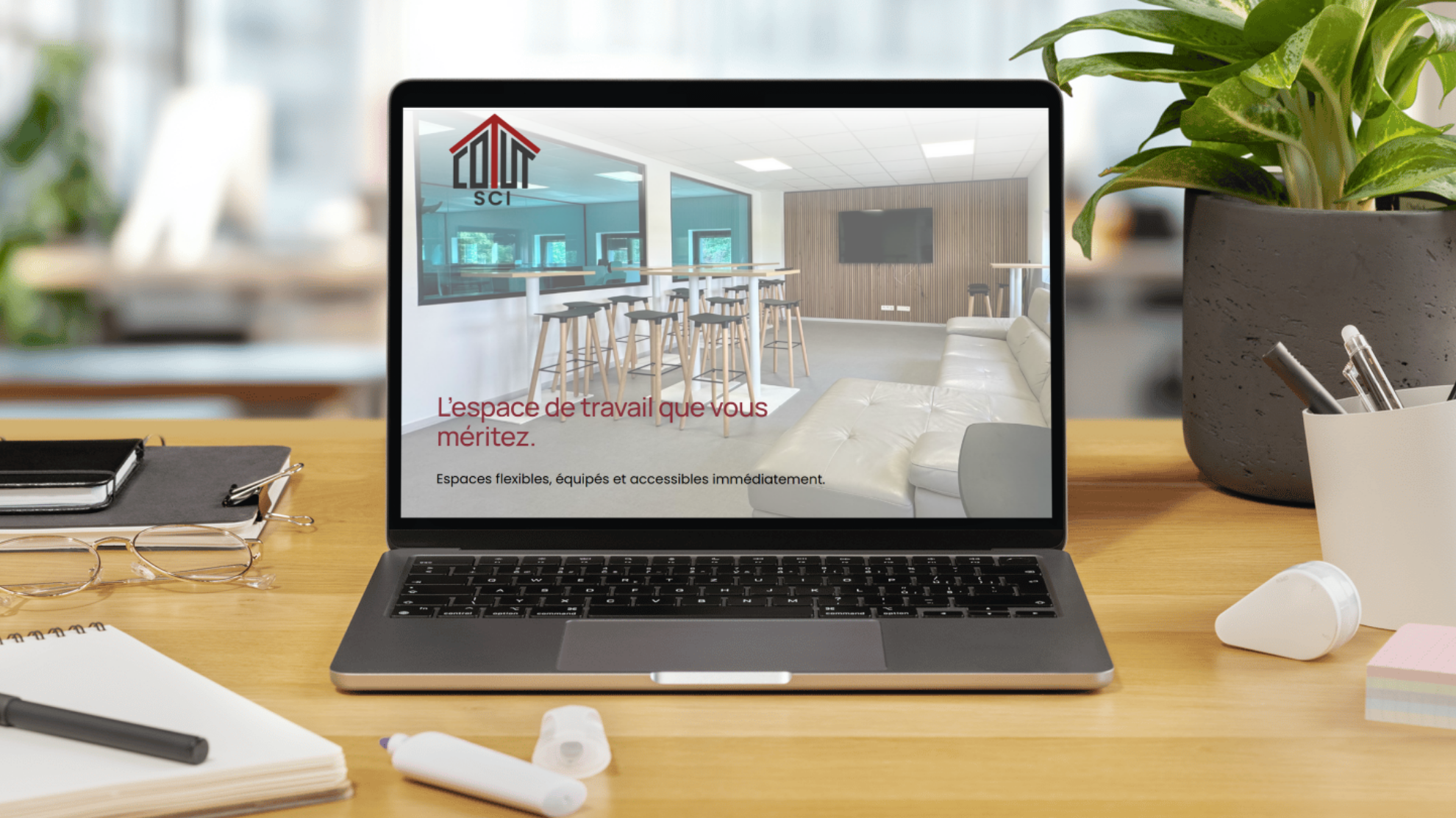 Un ordinateur portable sur un bureau en bois affiche une image d'un espace de coworking moderne et lumineux, avec un canapé blanc, des tables hautes et une grande télévision au mur. À côté de l'ordinateur, il y a des lunettes, un carnet, un stylo, de