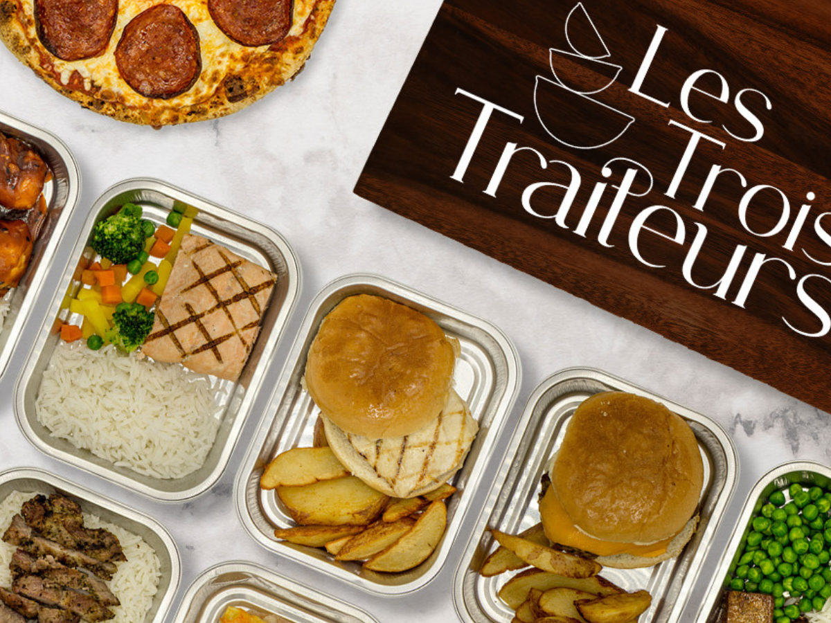 Assortiment de plats à emporter avec un pizza, des burgers, des légumes, des pommes de terre et un sign avec le texte "Les Trois Traiteurs" sur une planche en bois.