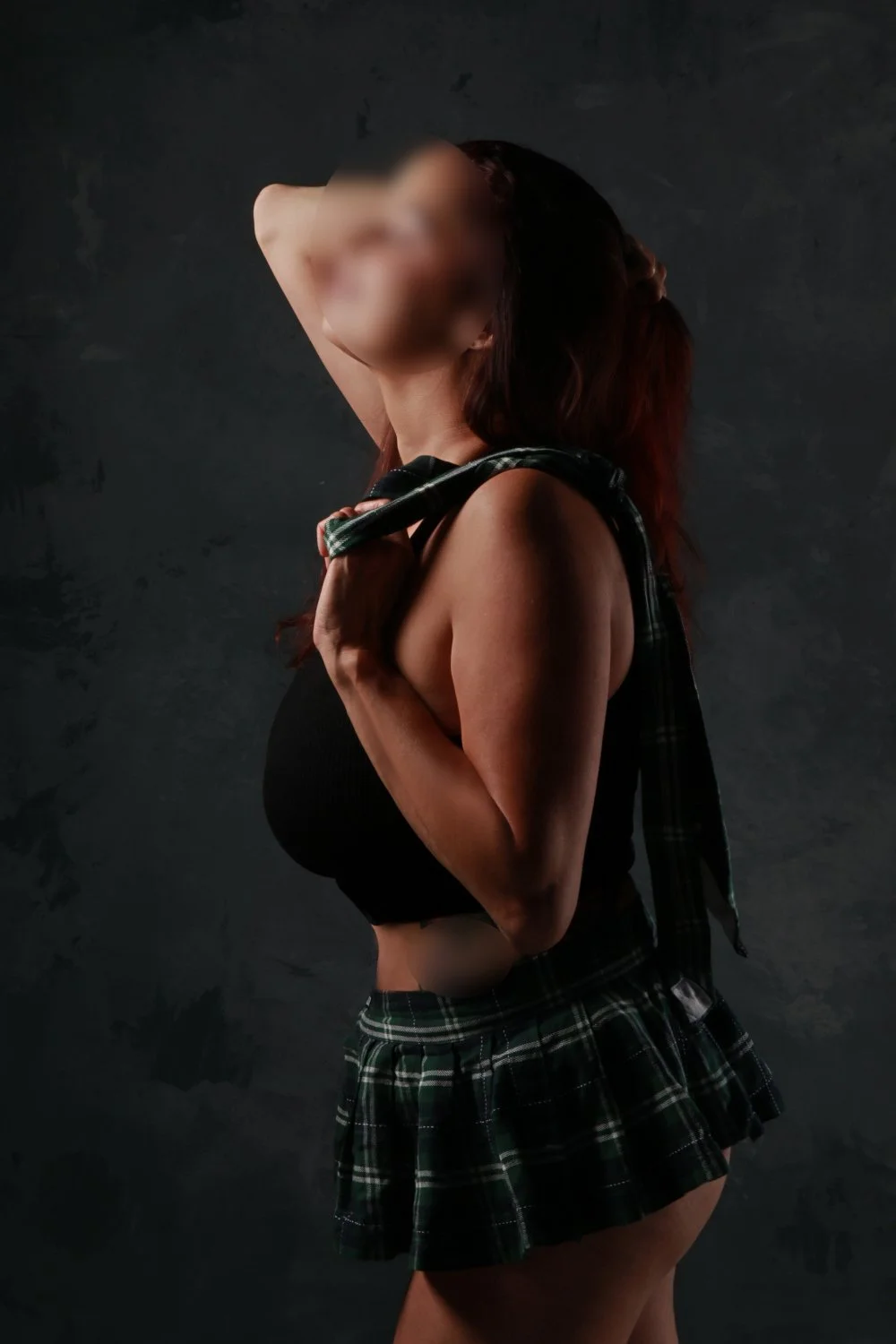 A woman with long hair and blurred face poses with one arm behind her head and the other holding a plaid shirt over her shoulder, wearing a black top and a plaid mini skirt, against a dark textured background.