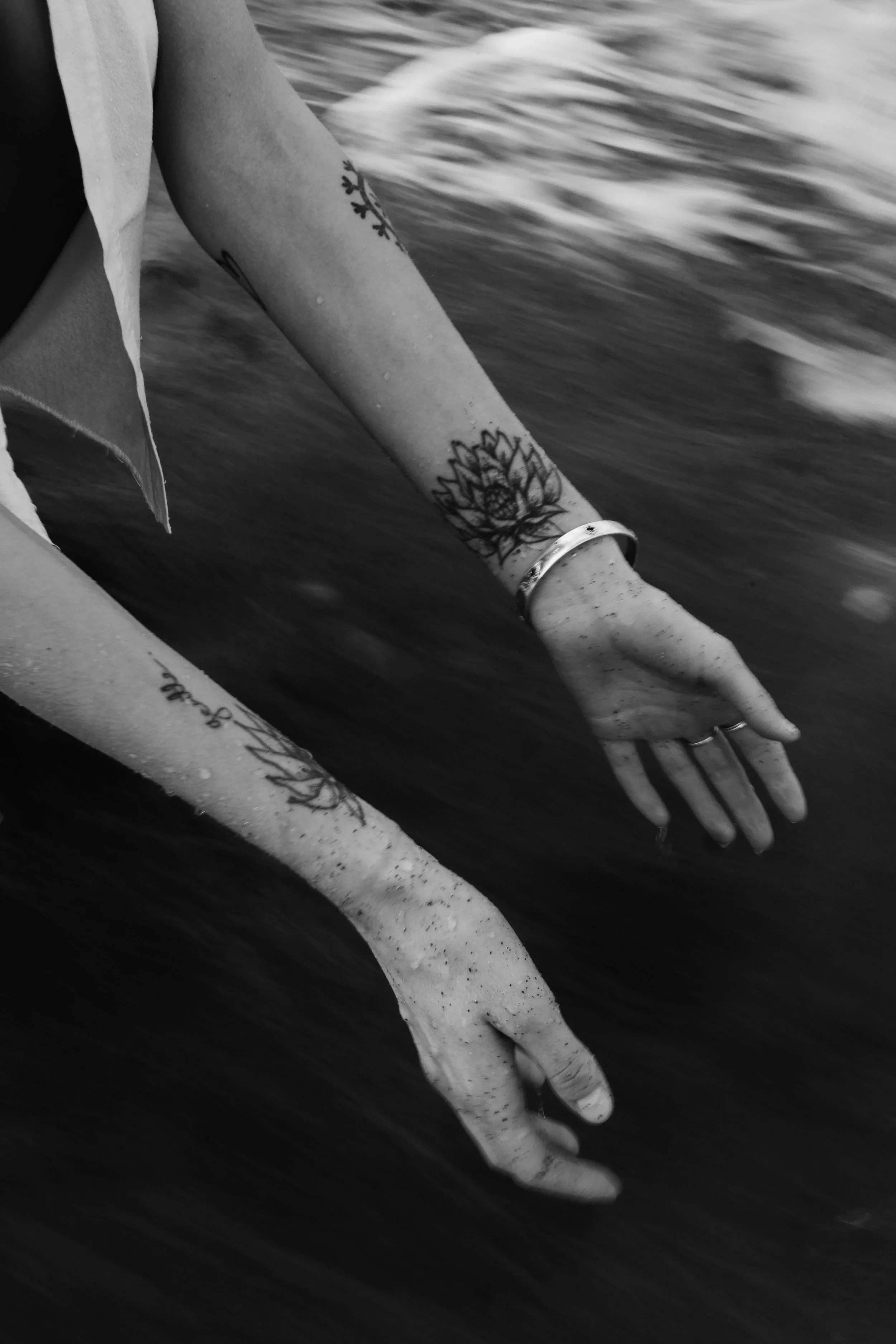 A person's arm and hand reaching into water, with visible tattoos on the arm, wearing a ring and a bracelet, in black and white.