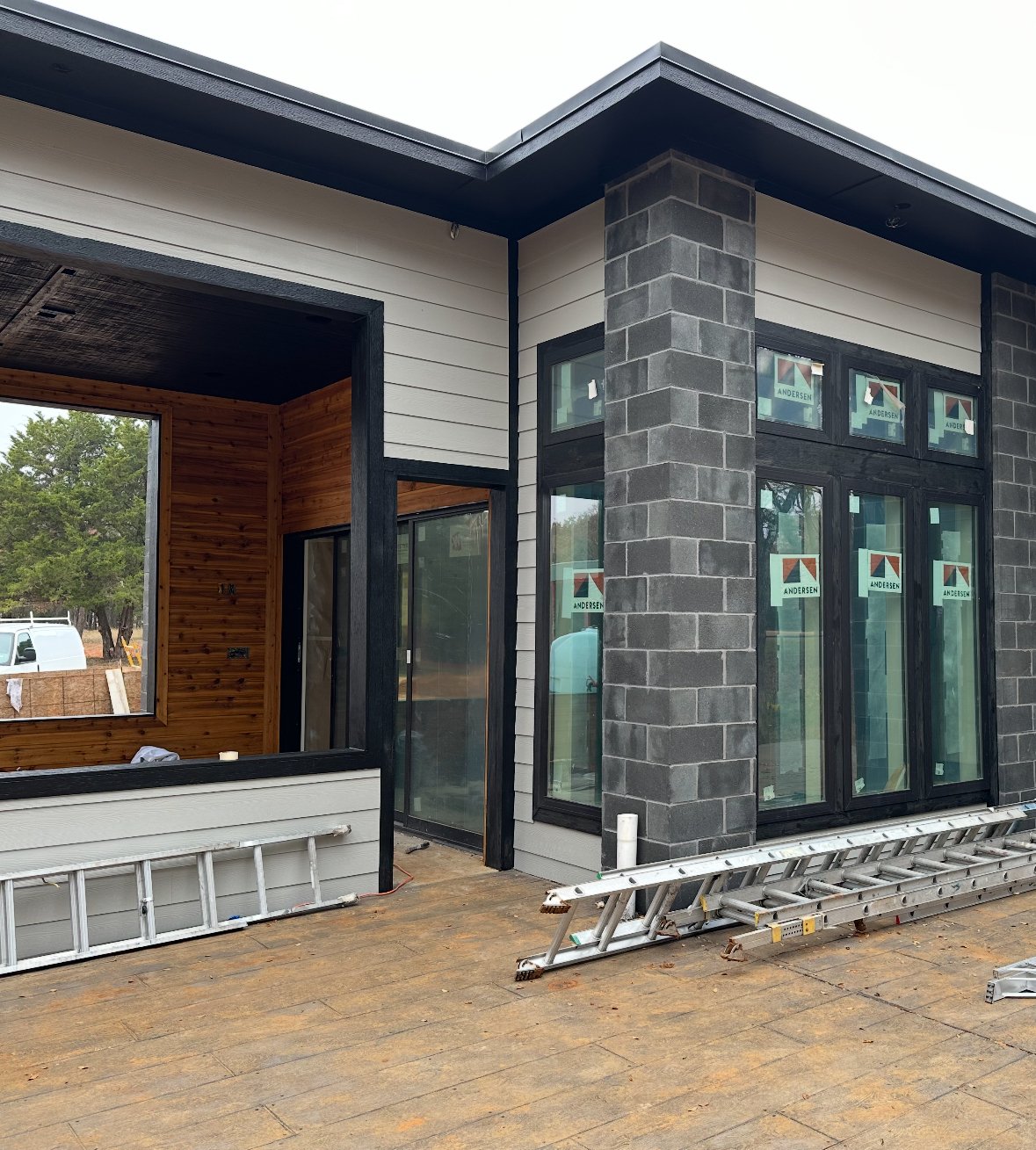 Exterior of a residential building under construction, with large windows covered in protective stickers, a gray brick column, and a wooden interior visible through an open section, with a ladder and materials on a wooden porch.