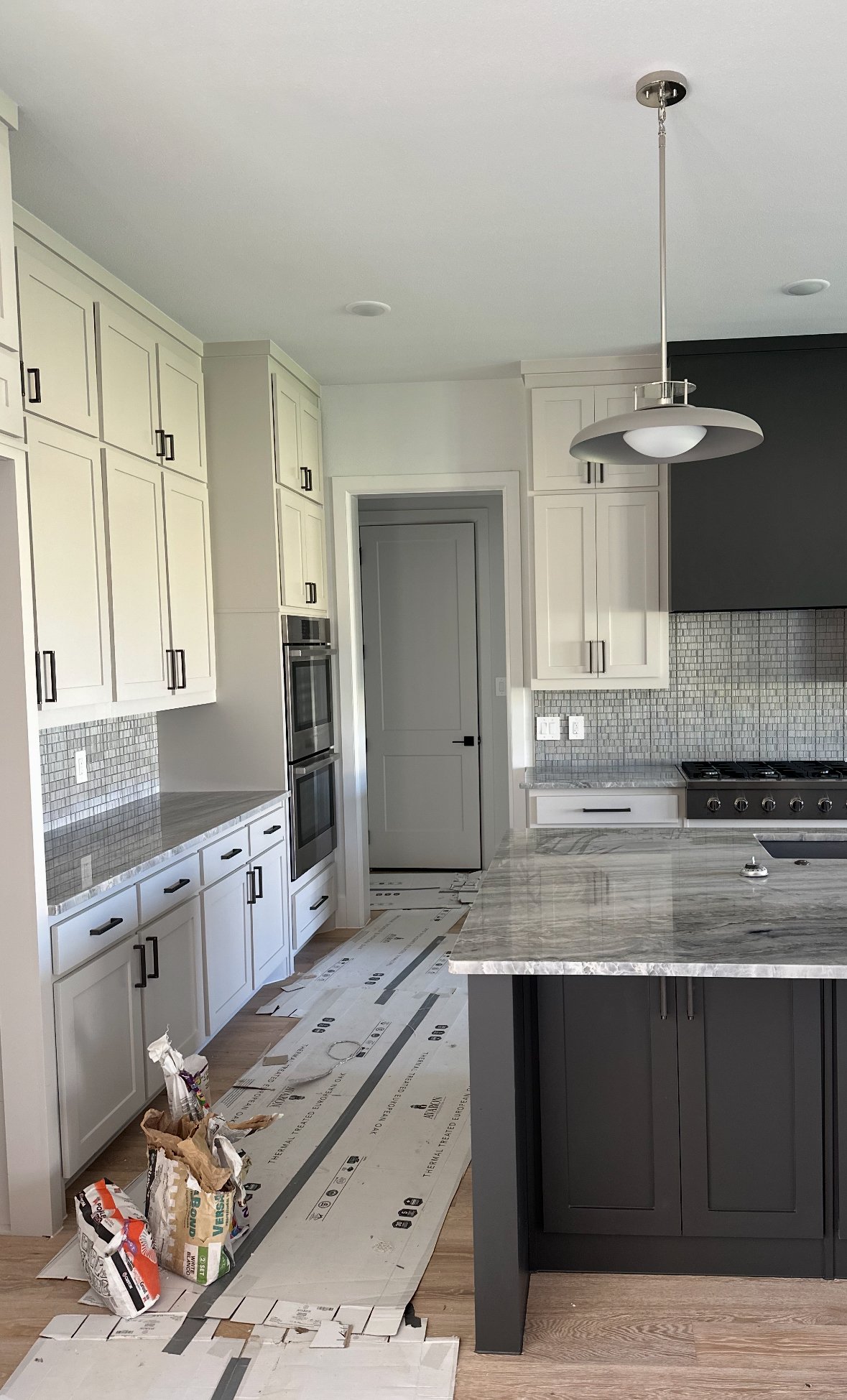 Newly renovated kitchen with white upper cabinets, dark lower cabinets, marble countertop island, gray backsplash tiles, and stainless steel appliances. Cardboard and bags of supplies are on the floor indicating ongoing work.