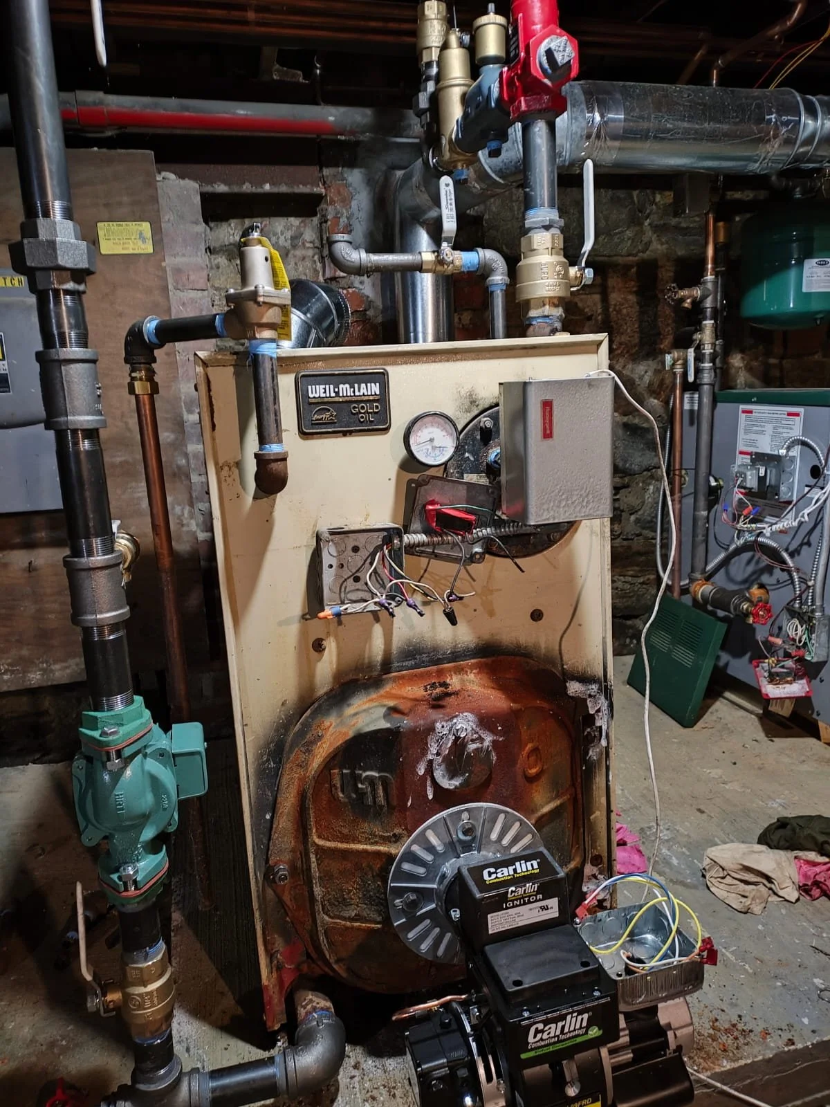 An old, rusty Weil-McLain Gold Oil boiler with various pipes, gauges, and wires connected, situated in a basement with exposed brick walls and clutter on the floor.