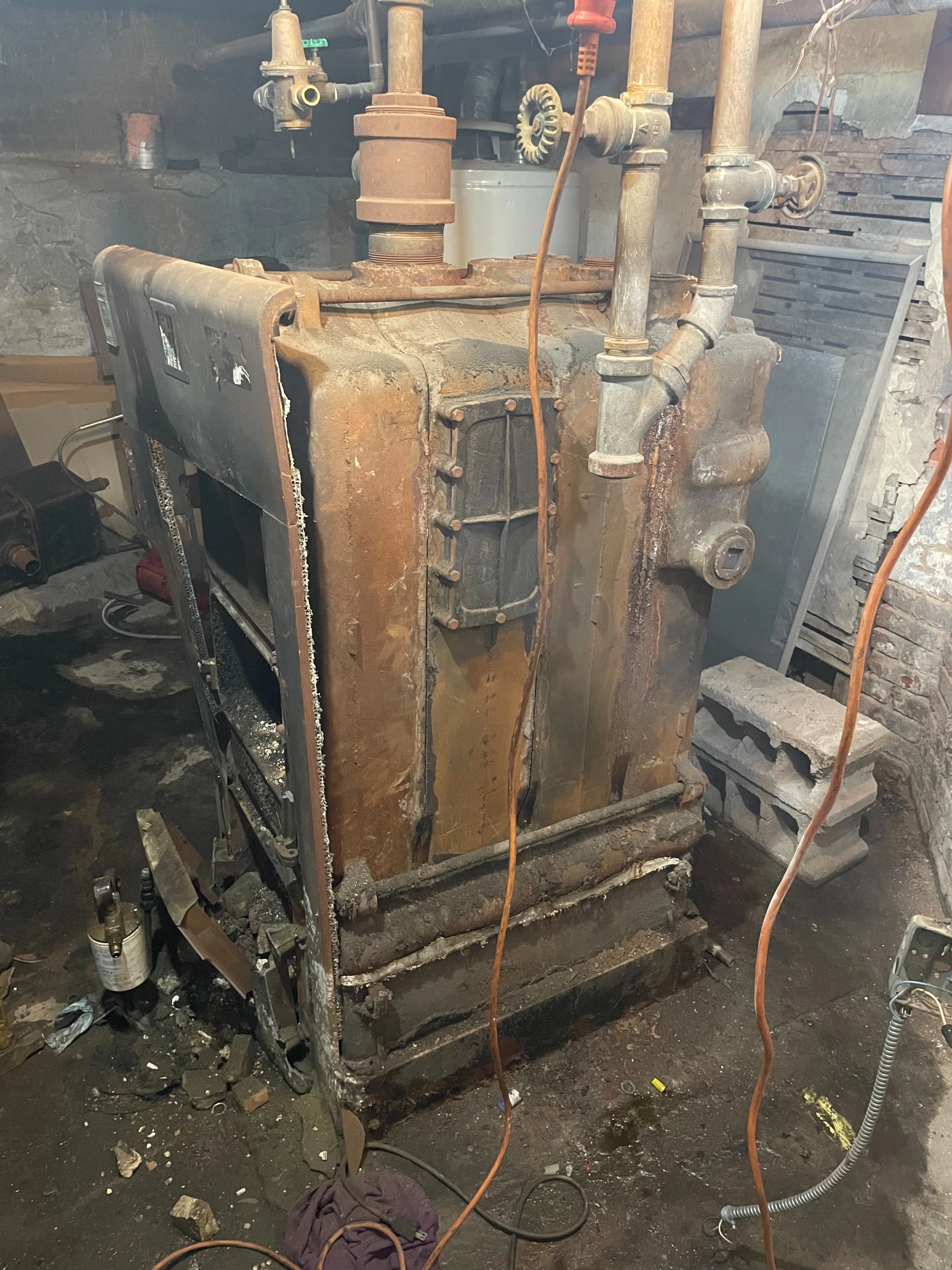 Old, rusty industrial boiler or furnace with visible pipes and valves, situated in a workshop or basement area.
