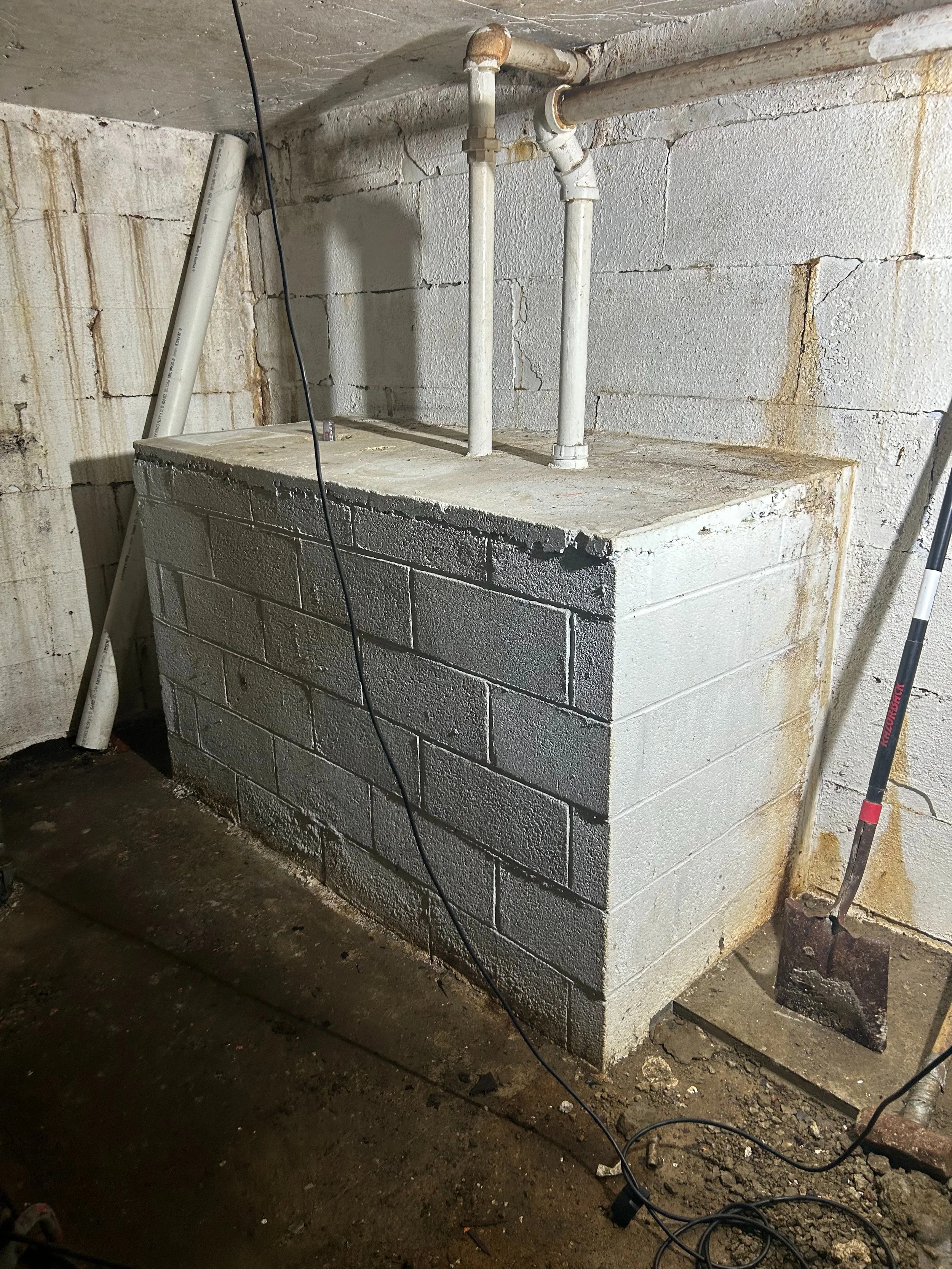 A concrete block foundation with plumbing pipes on an unfinished basement wall, a shovel leaning against the wall, and electrical cords laying on the ground.