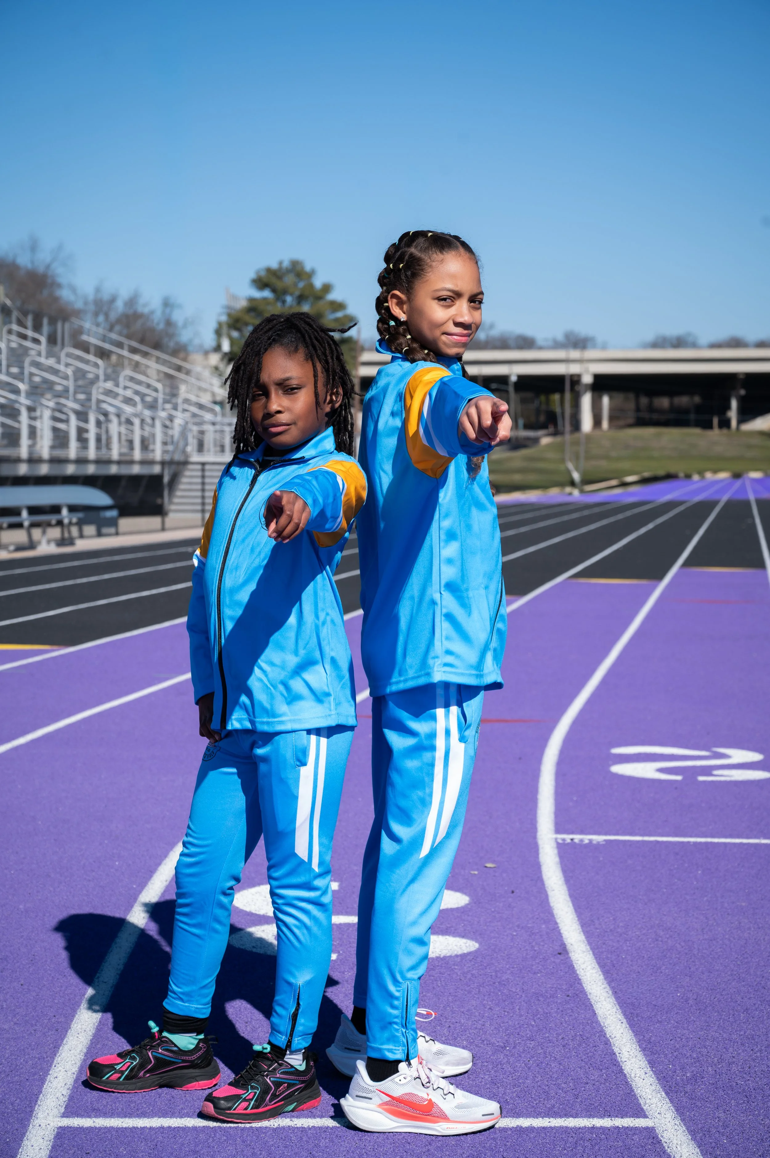 Two young girls in BHM Express track uniforms standing on a purple running track, pointing forward with confident expressions, against a clear blue sky and stadium bleachers.
