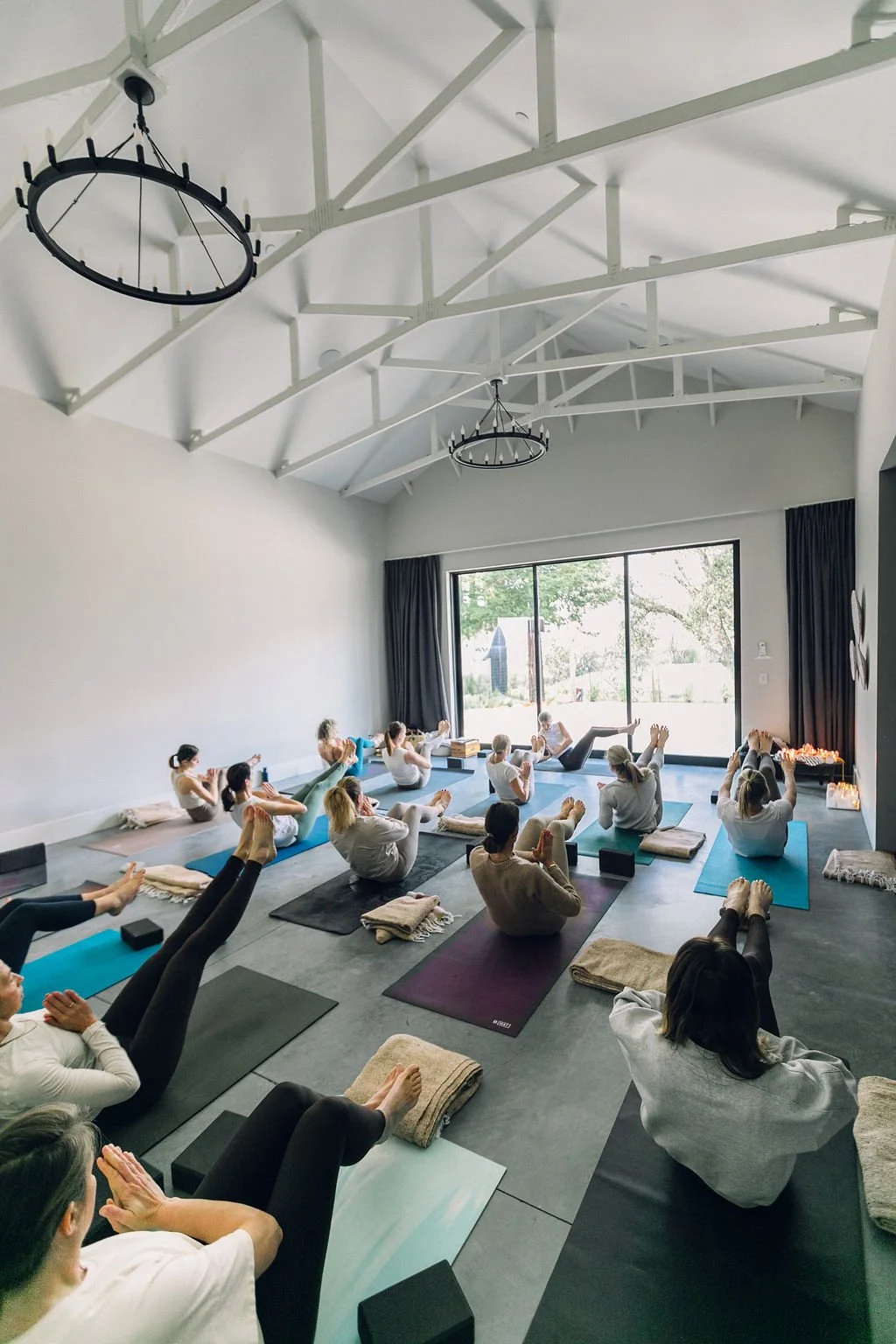 Indoor yoga class with participants practicing poses on mats, with natural light from large window, high white ceiling with exposed beams, black circular chandeliers, and candles on the side.