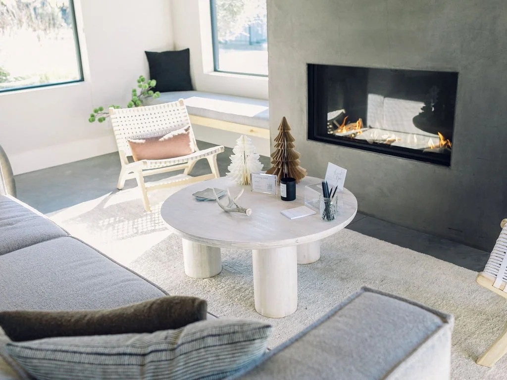 Modern living room with a white marble round coffee table, fireplace, light gray sofa, and white chair with cushions, decorated with artificial Christmas trees and holiday cards.