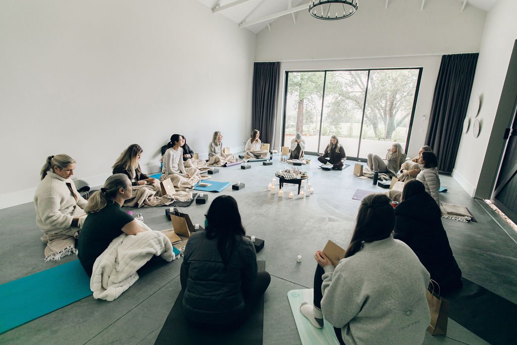 Group of people participating in a meditation or yoga session in a spacious, modern room with large window, curtains, and minimal decor.