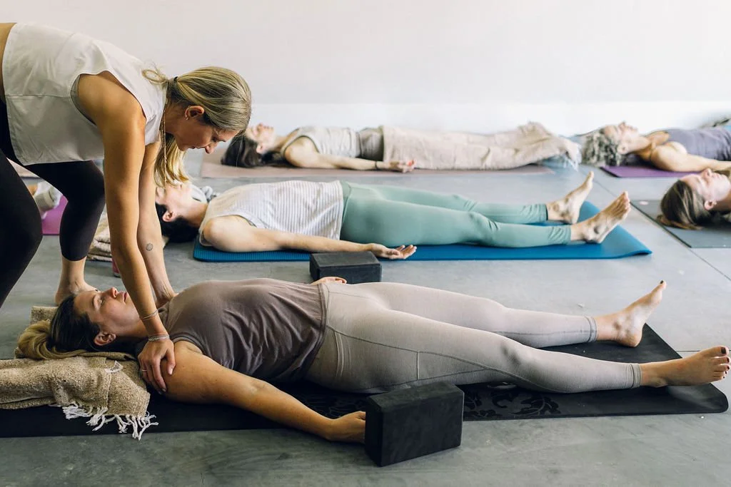 Women practicing yoga on mats in a studio, with an instructor assisting one woman.
