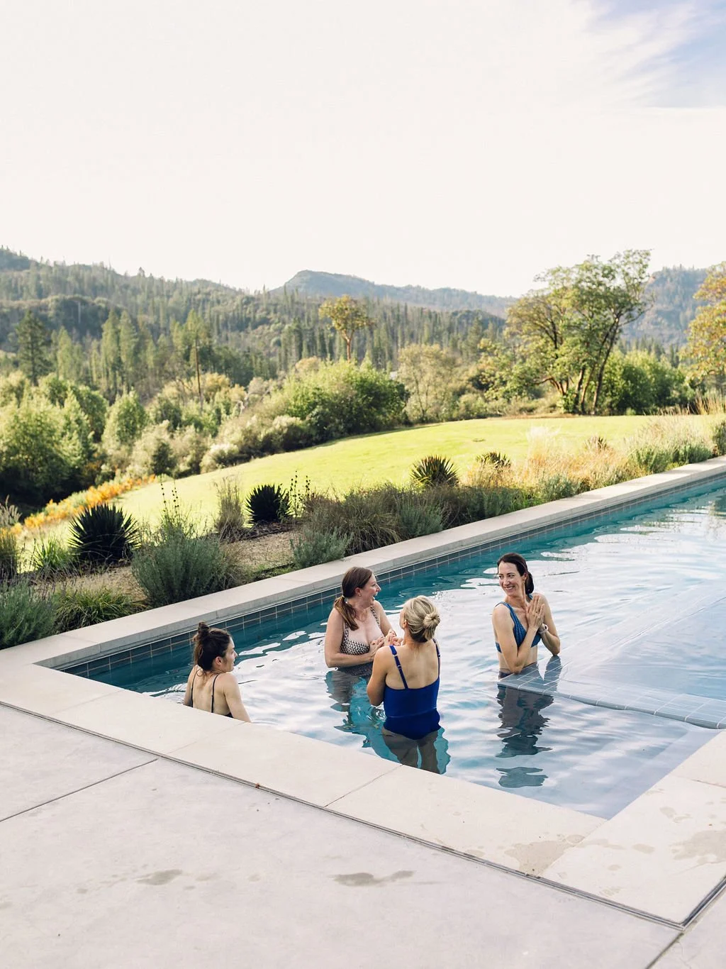 Four women are swimming and socializing in an outdoor pool with a scenic landscape of trees, rolling hills, and mountains in the background.
