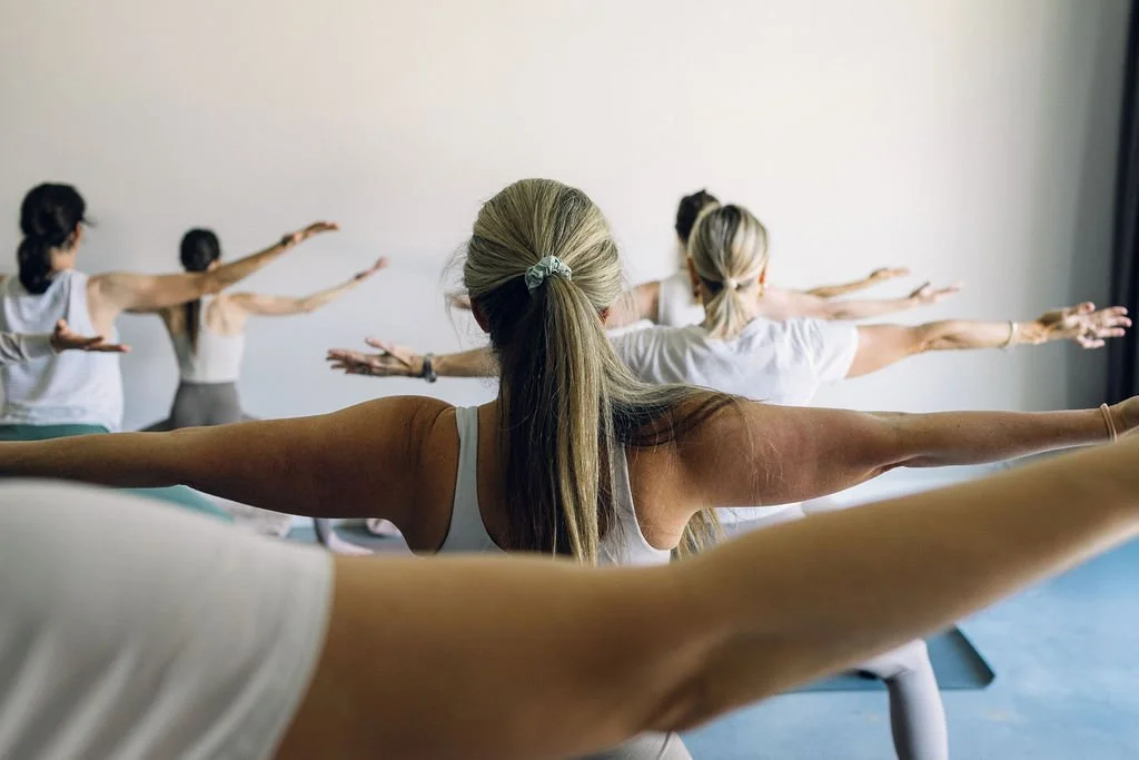 A group of women participating in a yoga class, extending their arms while doing a stretch exercise in a well-lit room.