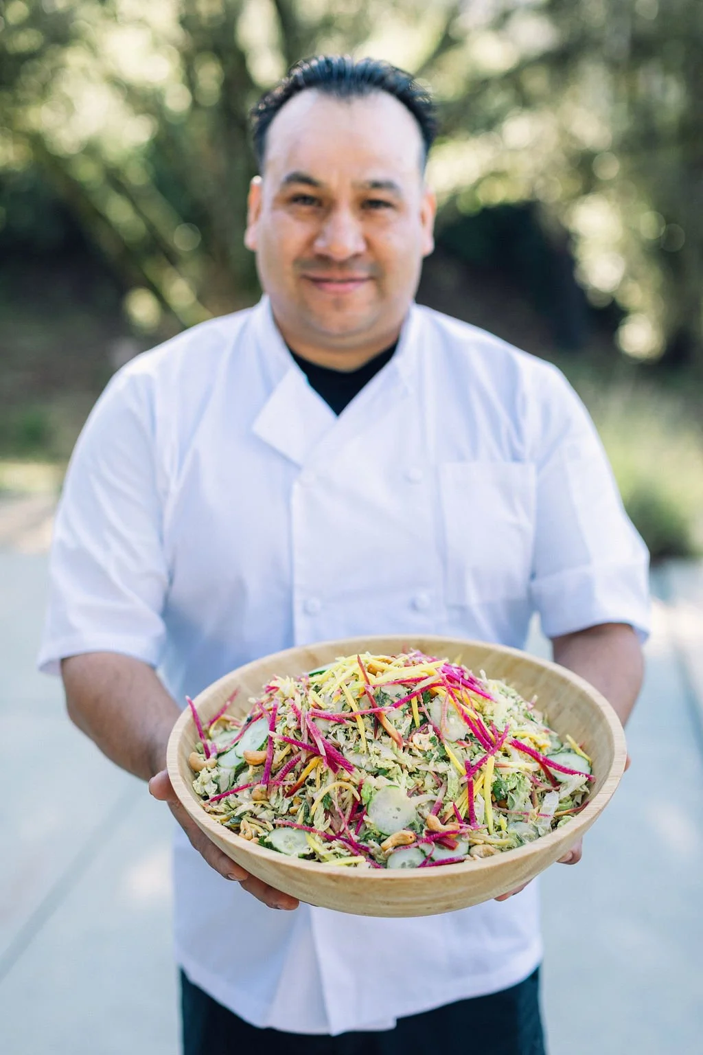 Chef in white uniform holding a bowl of colorful salad outdoors with trees in the background.