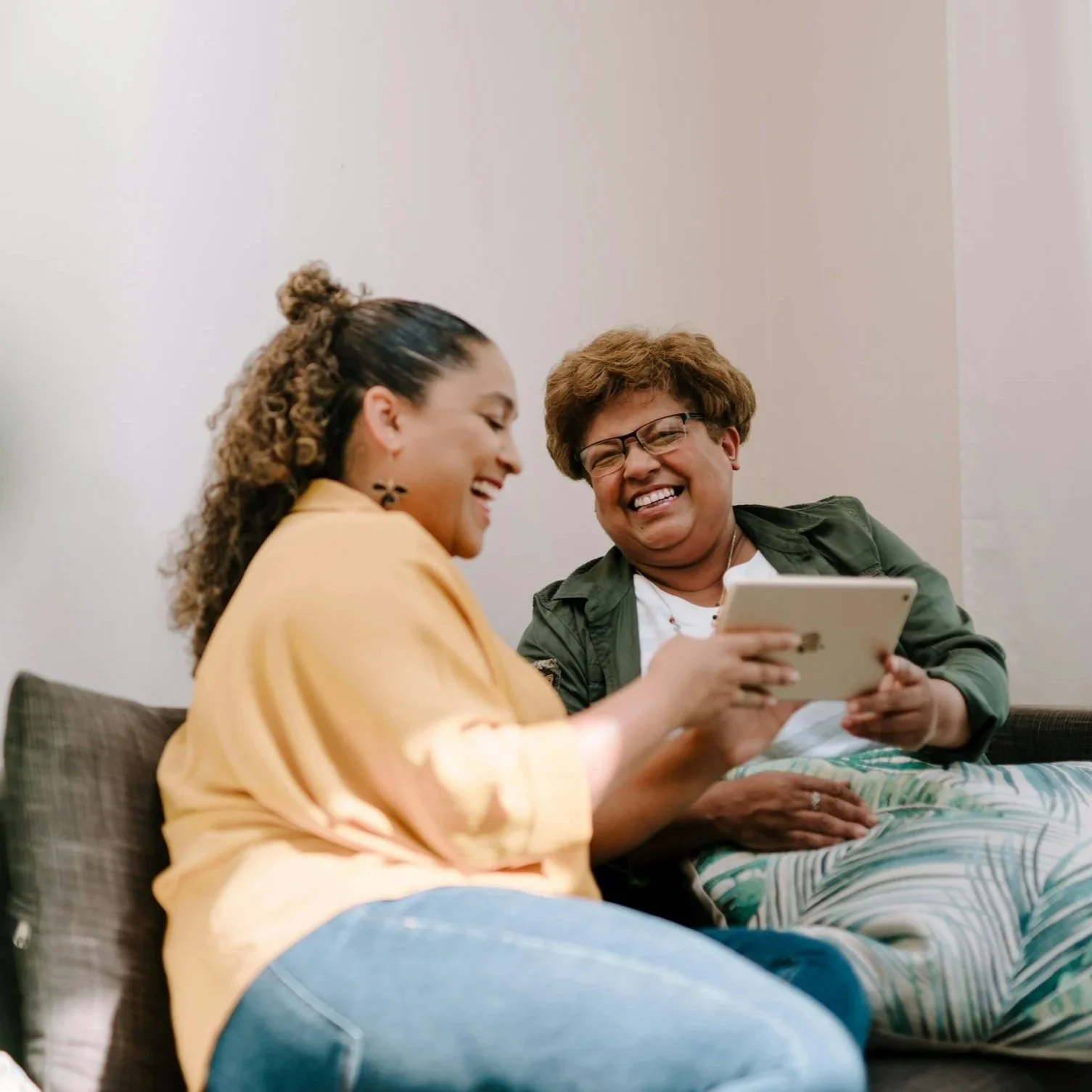 Two women sitting on a couch, laughing and looking at a tablet together, enjoying a joyful moment.